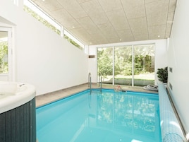 An indoor swimming pool with a hot tub, large windows, and a view of greenery outside.