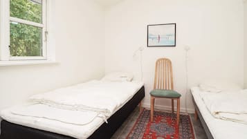 Ein kleines, ordentliches Zimmer mit zwei Einzelbetten, einem Sessel, einem Teppich und einem Fenster mit Blick auf Bäume.