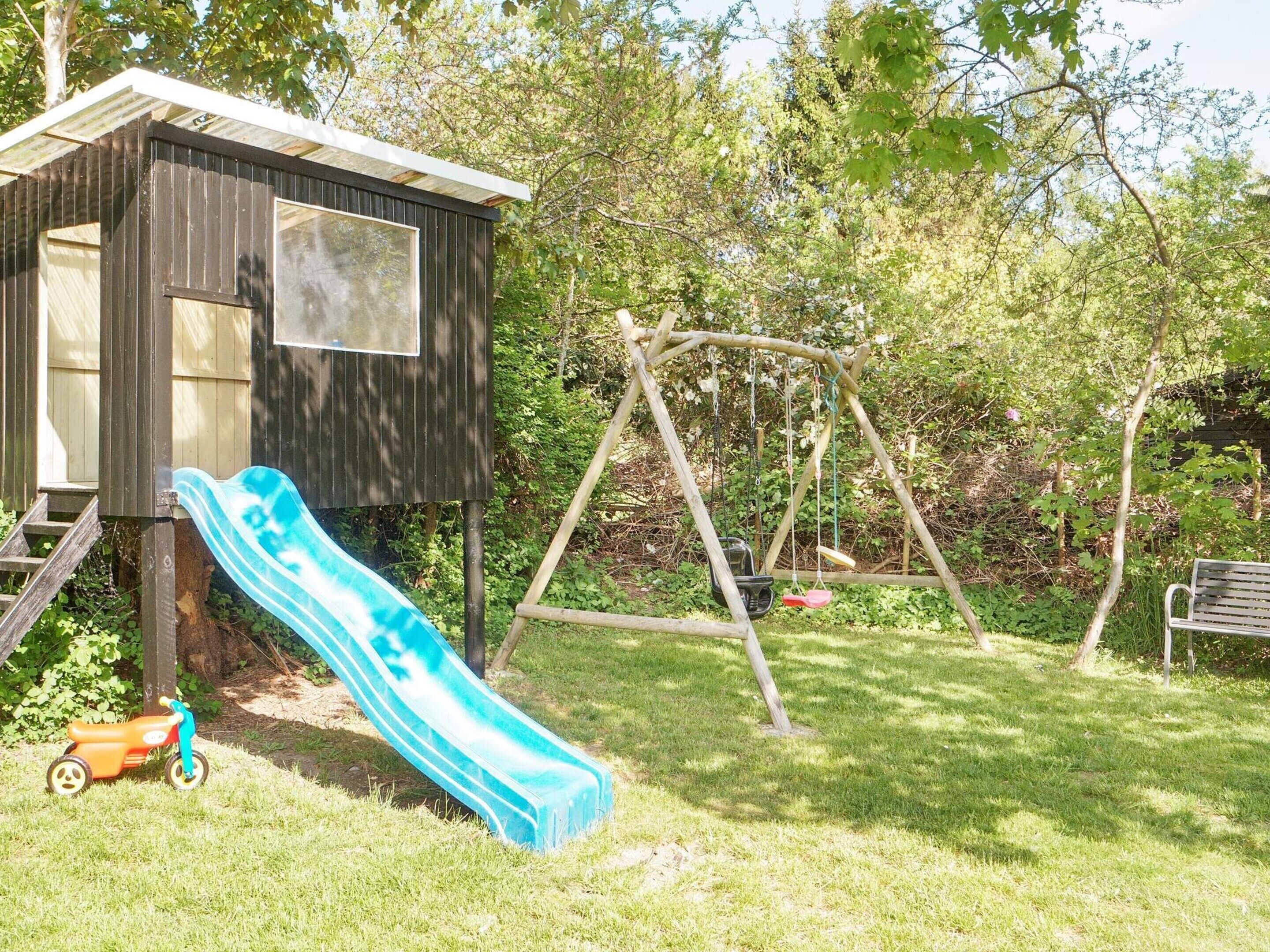 A wooden playhouse with a slide and a swing set in a grassy yard.