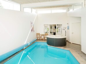 A modern indoor swimming pool with a hot tub, wooden chairs, and a kitchen area visible through glass doors.
