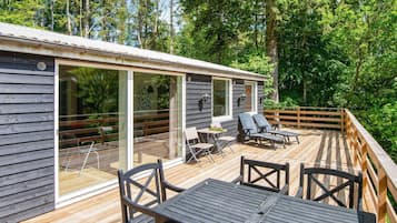 Una terraza de madera con mobiliario exterior, una puerta de entrada de vidrio y vistas a un área boscosa.
