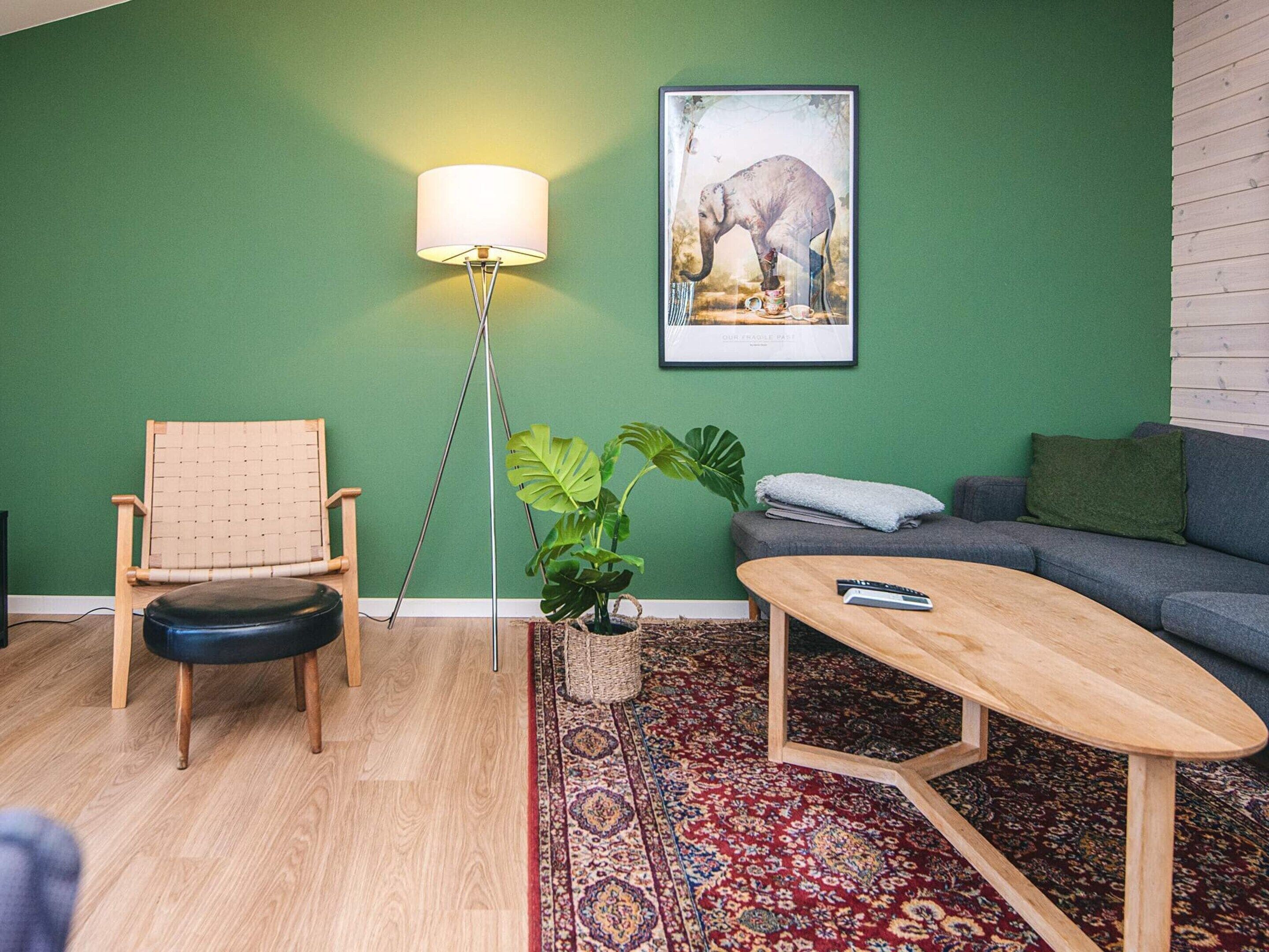 Una sala de estar con pared verde, suelo de madera, un sofá, un sillón, una mesita de centro, una lámpara de pie y un cuadro con una imagen de un elefante.
