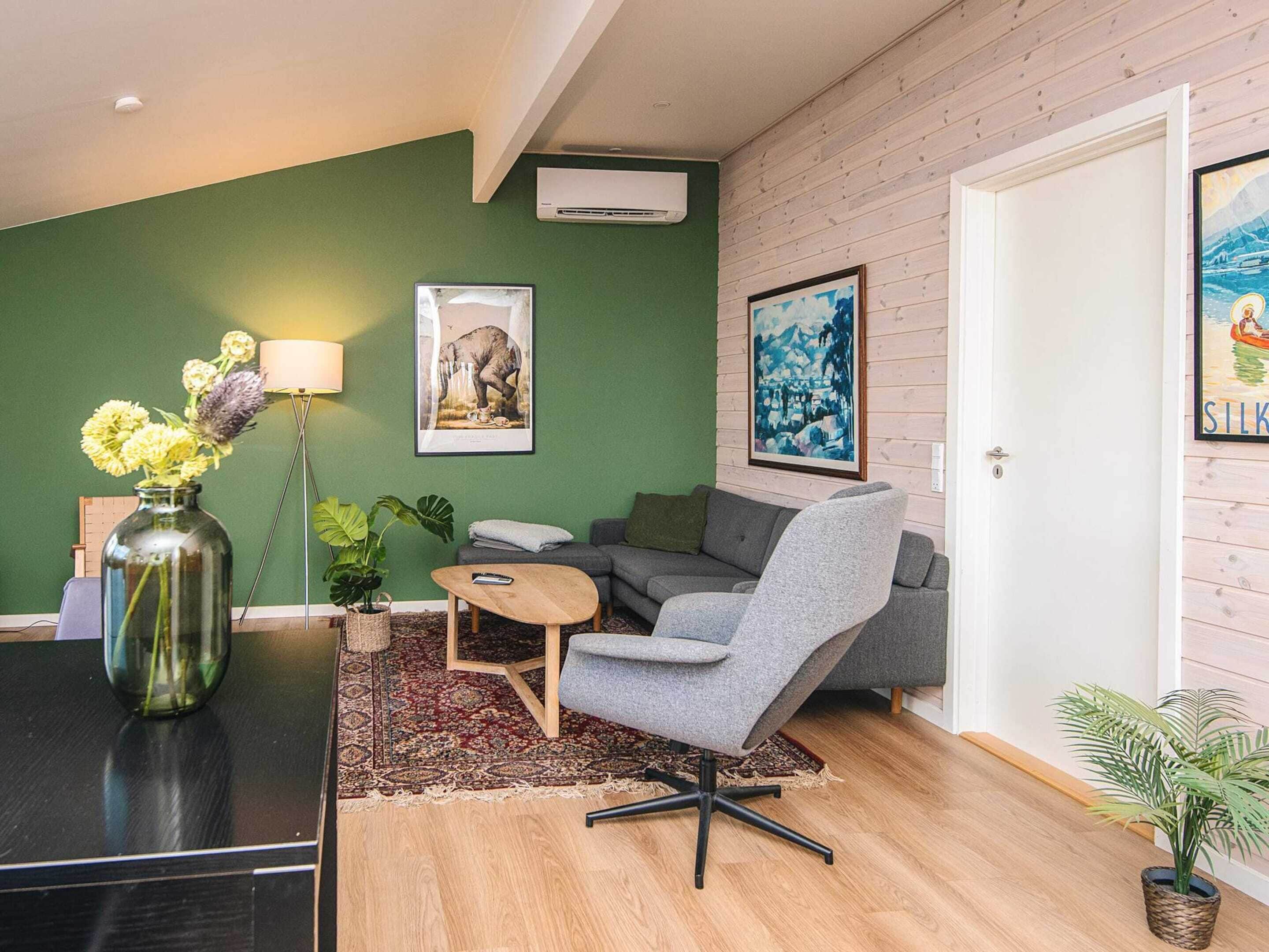 Una sala de estar moderna con una pared verde, un sofá gris, un sillón gris, una mesa de centro de madera y una alfombra estampada.