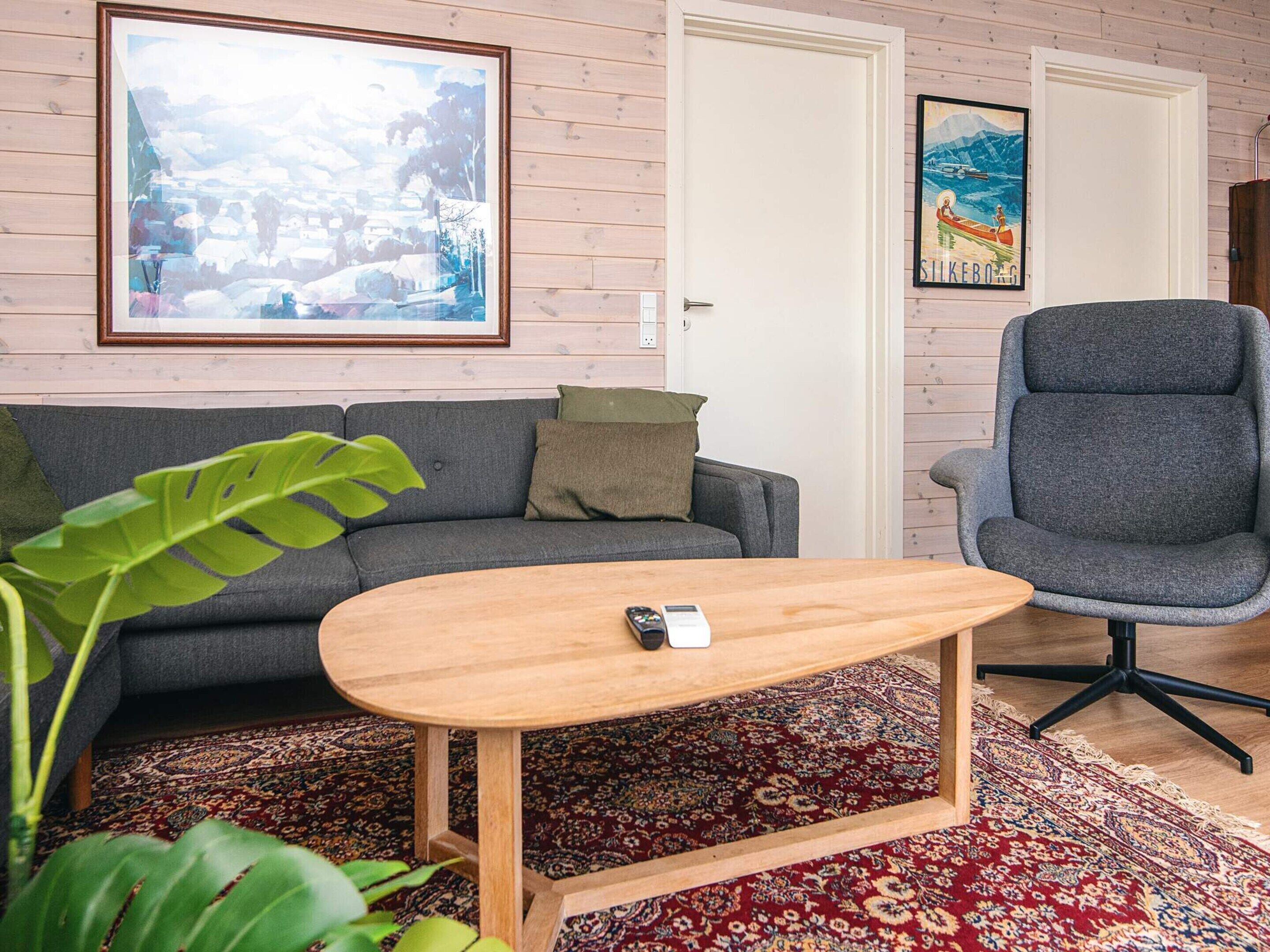 Un salon avec un canapé gris, une table basse en bois, un fauteuil de bureau gris, un grand tableau encadré et un tapis à motifs.