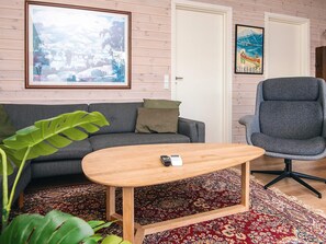Ein Wohnzimmer mit grauem Sofa, einem Holztisch, einem grauen Bürostuhl, einem großen gerahmten Bild und einem gemusterten Teppich.