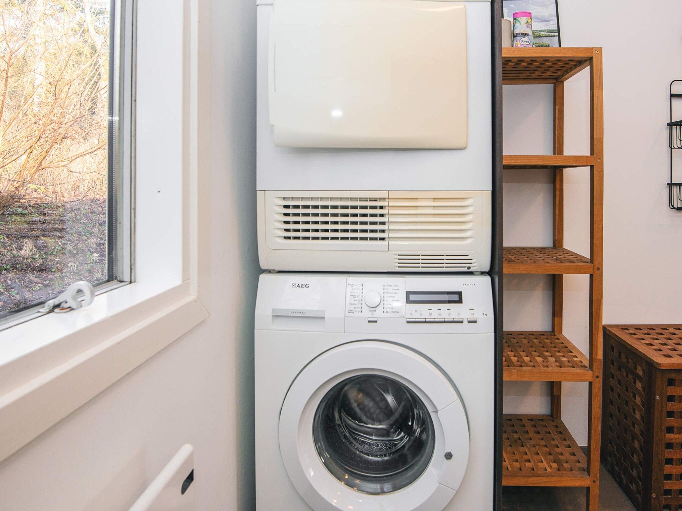 A front-loading washing machine with a control panel, a dryer above it, and a wooden shelf unit to the right.