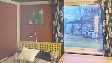 A bedroom with a bed, a bedside table, a window with curtains, and a view of a small house outside.