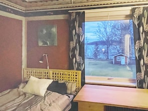 A bedroom with a bed, a bedside table, a window with curtains, and a view of a small house outside. - 6 Person Holiday Home in Amal (Åmål)