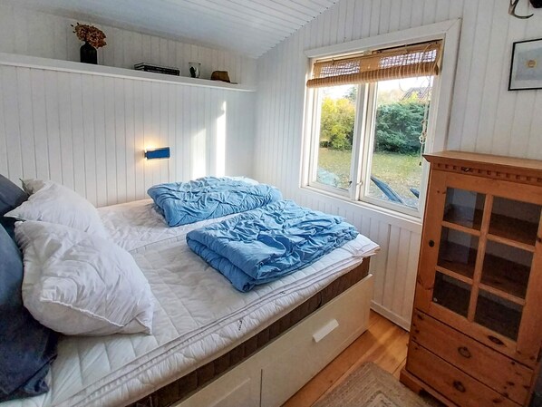 Ein Schlafzimmer mit einem Bett, einer Holzkommode und einem Fenster mit Blick auf Grünflächen.