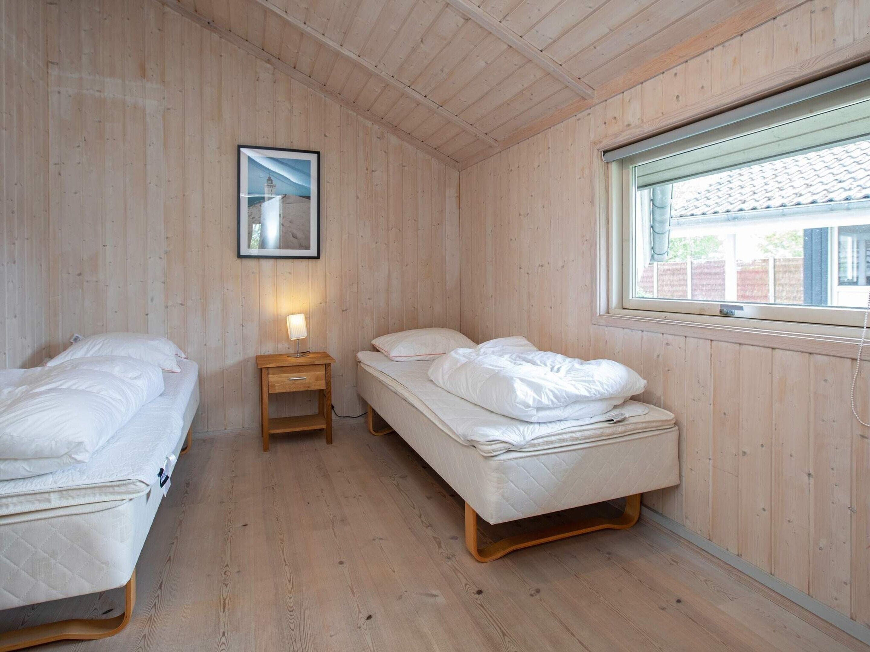 Een kamer met houten lambrisering, twee bedden, een nachtkastje met lamp en een raam met uitzicht op een gebouw.