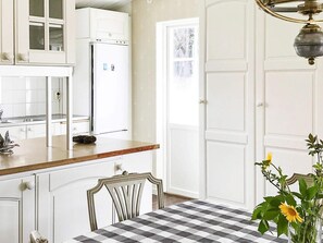 Une cuisine avec des meubles blancs, un réfrigérateur, une table à manger recouverte d’une nappe à carreaux et un vase rempli de fleurs.