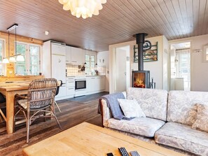 A cozy living room with a sofa, a wooden coffee table, and a dining area with a wooden table and wicker chairs. A fireplace is visible in the background. - 4 Person Holiday Home in Oksbol (Oksbol)
