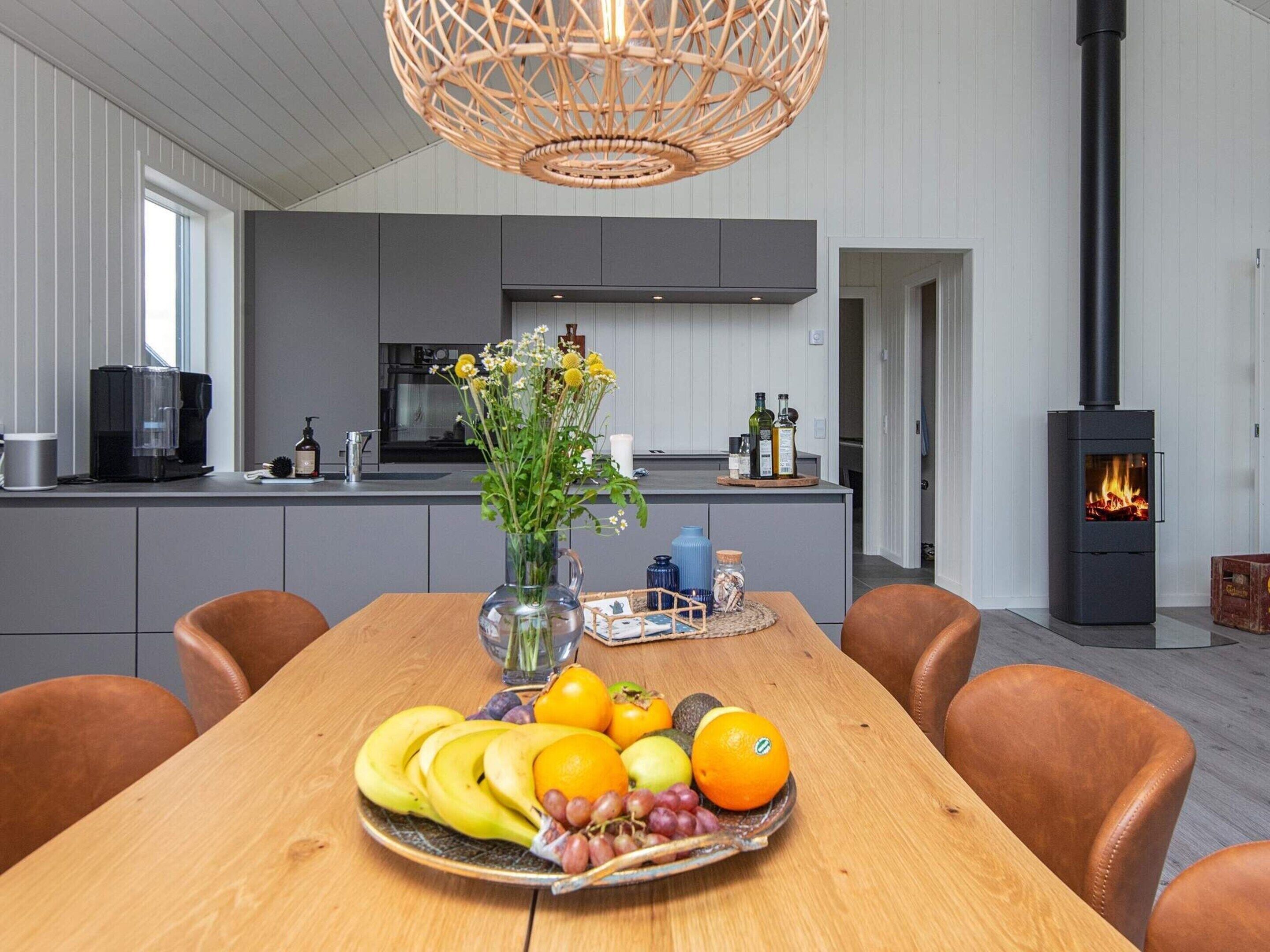 A modern kitchen with a central island, a dining table with fruit, and a wood-burning stove.