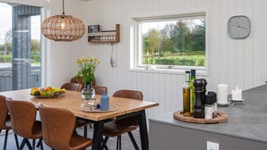 En matplats med ett träbord, bruna stolar, en hängande lampa, ett fönster med utsikt över grönskan och en klocka på väggen.
