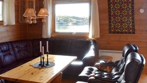 Interior acolhedor de uma cabana, com uma mesa de madeira, sofás de couro e vista para o exterior através de uma janela.
