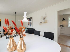 Un espace repas moderne avec une table blanche, deux vases contenant des plantes séchées, un luminaire suspendu blanc et une cuisine en arrière-plan.