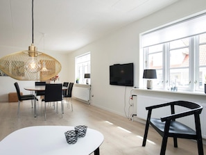 A modern living room with a dining area, a TV mounted on the wall, and a large window providing natural light.