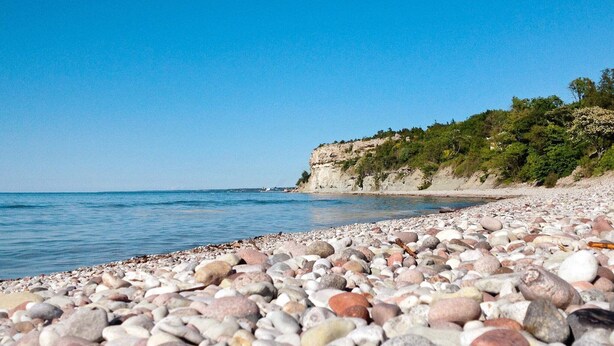 A pebbled beach with a clear blue sea and a rocky cliff with vegetation. - 6 Person Holiday Home in Visby-by Traum (Visby)