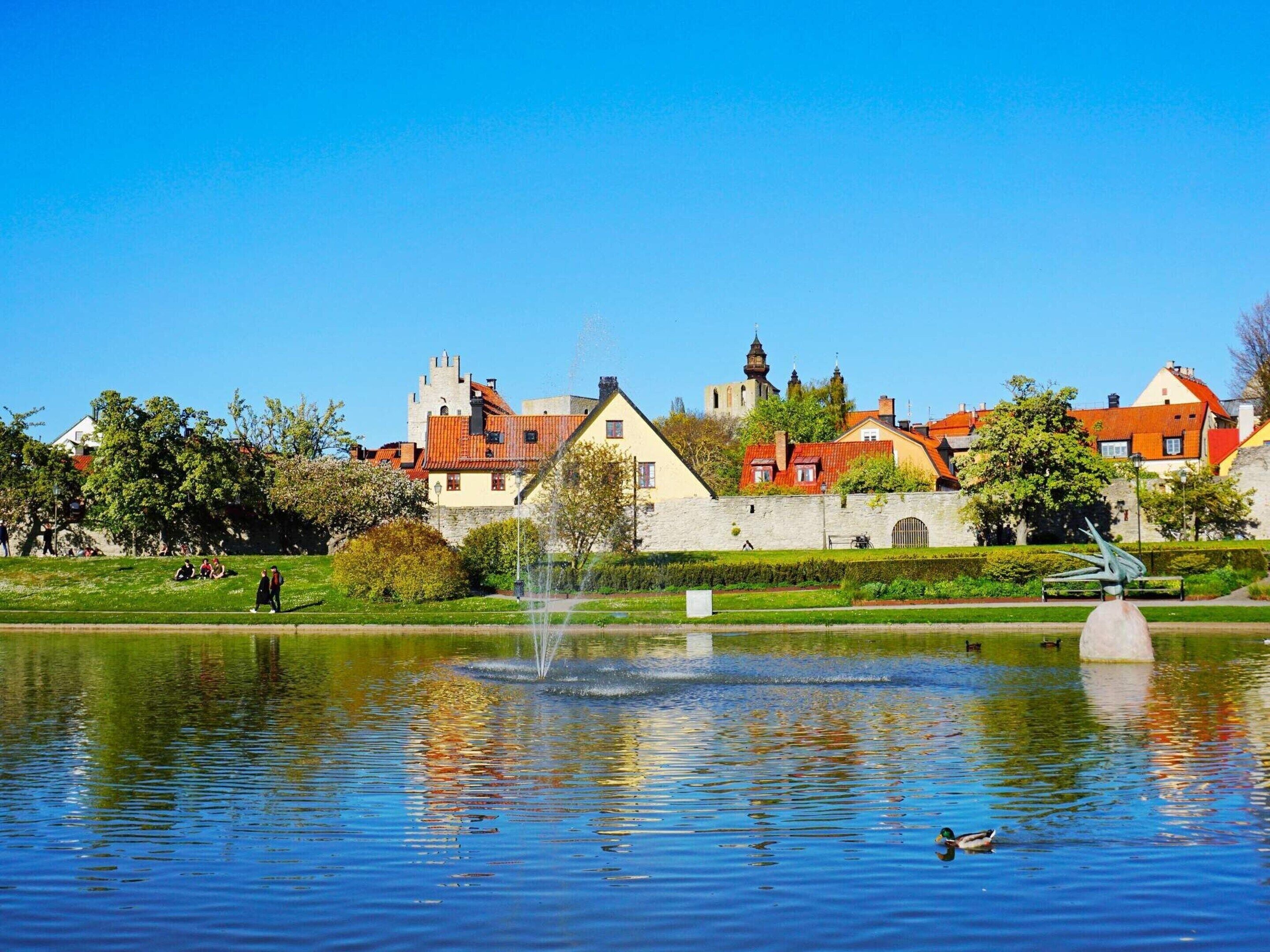 A serene park with a fountain, a duck swimming, and historic buildings in the background.