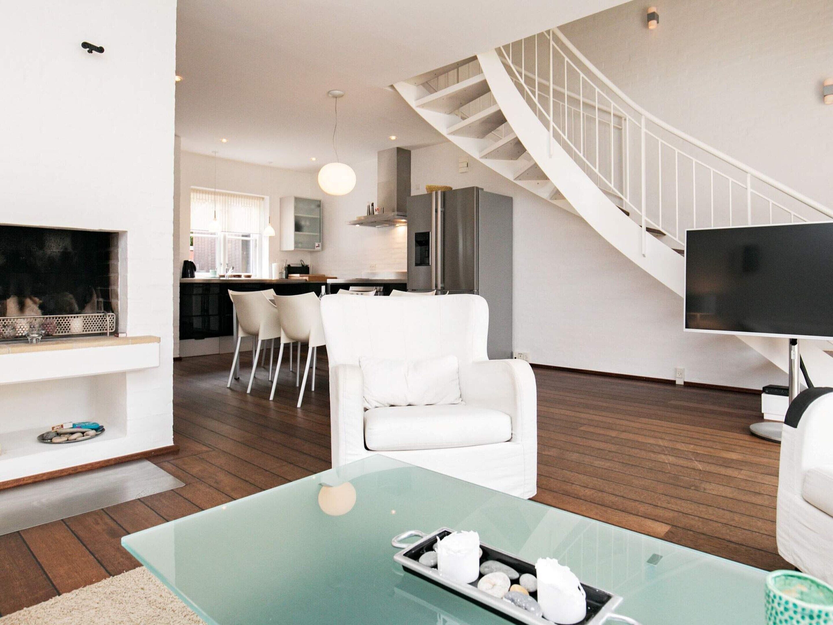 A modern living room with a glass coffee table, white sofas, a fireplace, and a staircase leading to an upper level.