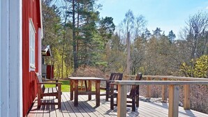 Una terraza de madera con un edificio rojo, mobiliario para exteriores y un paisaje boscoso de fondo.