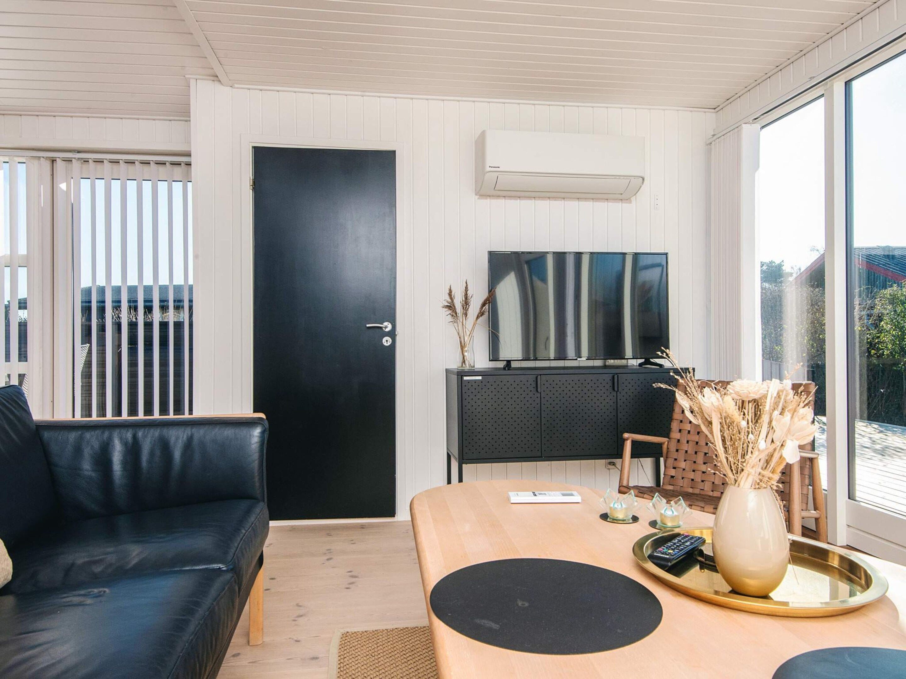 A modern living room with a black leather sofa, a wooden dining table, a flat-screen TV mounted on the wall, and a large window providing a view of the outdoors.