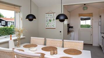 A dining area with a white table, wicker chairs, black pendant lights, and a botanical print on the wall.
