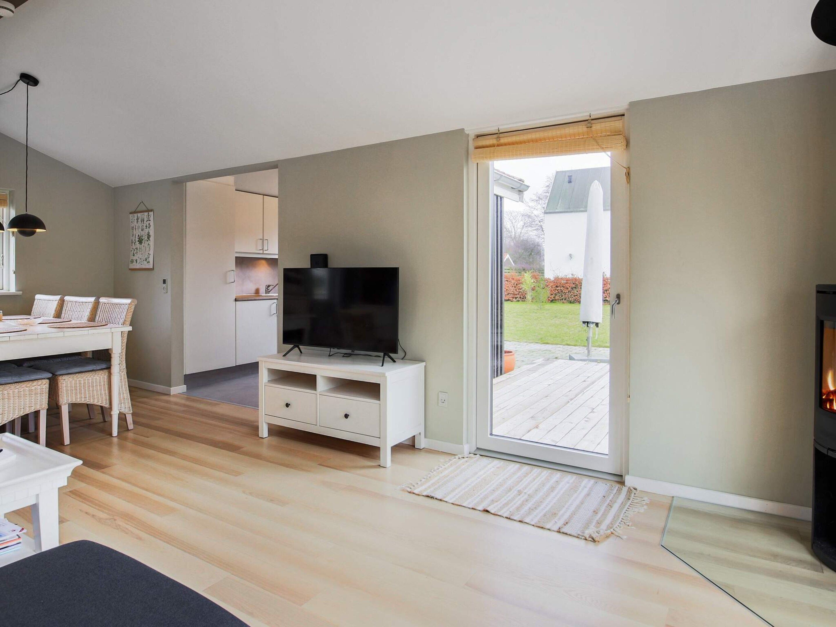 A modern living room with a flat-screen TV on a stand, a dining area with a white table and wicker chairs, and a sliding glass door leading to a wooden deck.