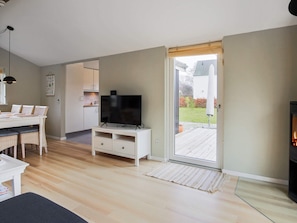 A modern living room with a flat-screen TV on a stand, a dining area with a white table and wicker chairs, and a sliding glass door leading to a wooden deck. - 6 Person Holiday Home in Vaeggerlose (Vaeggerlose)
