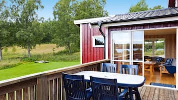A wooden deck with a table and chairs, a covered seating area with a view of the outdoors, and a red building with a window.