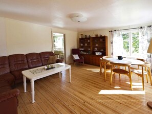 A living room with a brown leather sofa, a white coffee table, and a wooden dining table with chairs. There is a bookshelf filled with books and decorative items.
