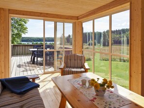 A room with a large window, a wooden deck outside, a sofa, a table with a vase of flowers, and a chair.