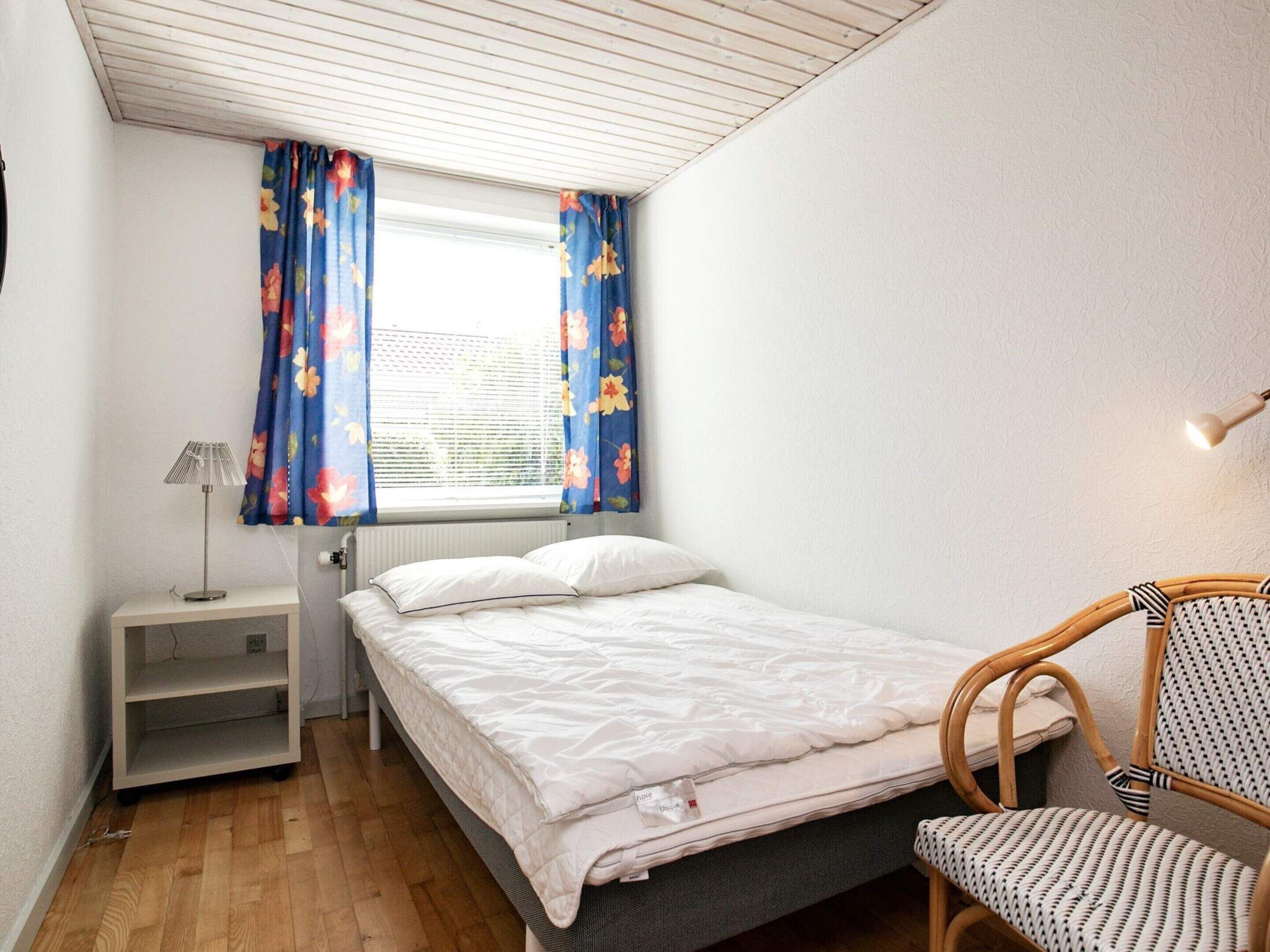 A small, neatly arranged bedroom with a bed, a chair, a bedside table, and a window with floral curtains.