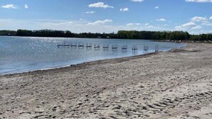 Una playa arenosa con un cuerpo de agua tranquilo, una fila de postes de madera y una orilla boscosa bajo un cielo despejado.