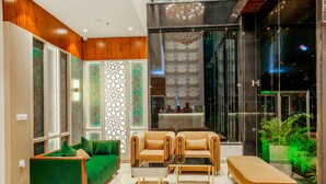 A modern lobby with a green sofa, a beige sofa, and a bench. There is a glass partition with a geometric pattern and a chandelier. - HOTEL EUPHORIA GREEN (Amritsar)