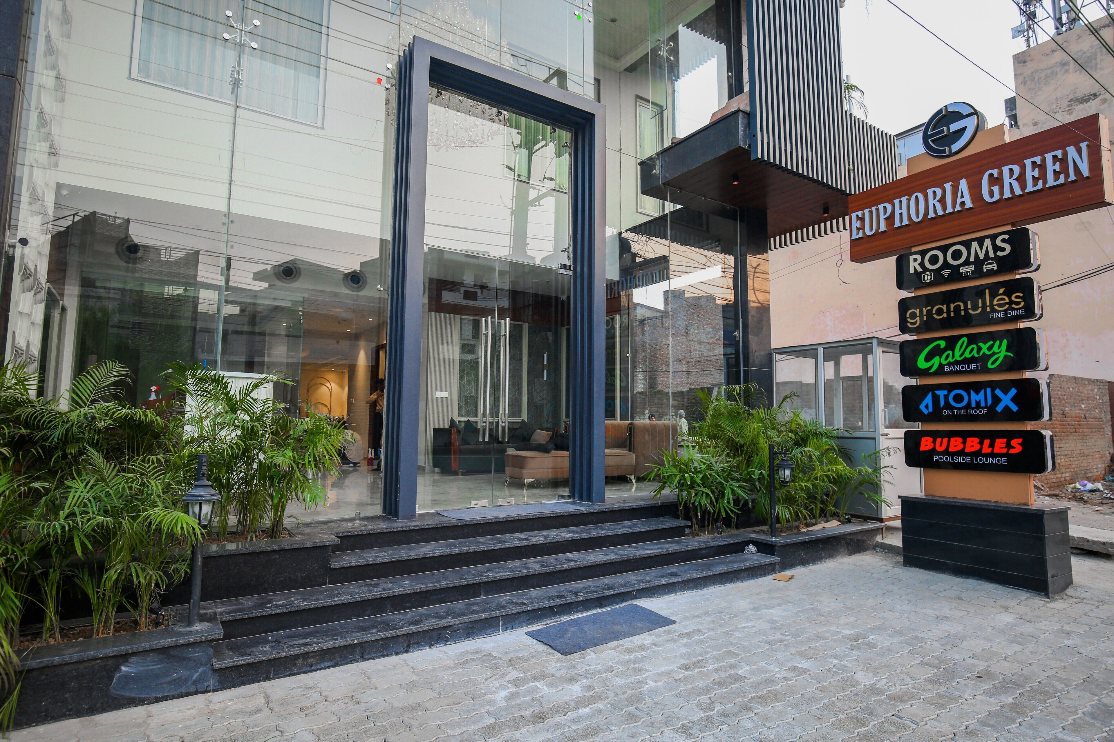 En hotellentrén med en modern glasfasad, en trappa som leder upp till entrén och skyltar som visar vägen till olika faciliteter, inklusive rum, en finrestaurang, en bankettsal, en bar på takterrassen och en lounge vid poolen.