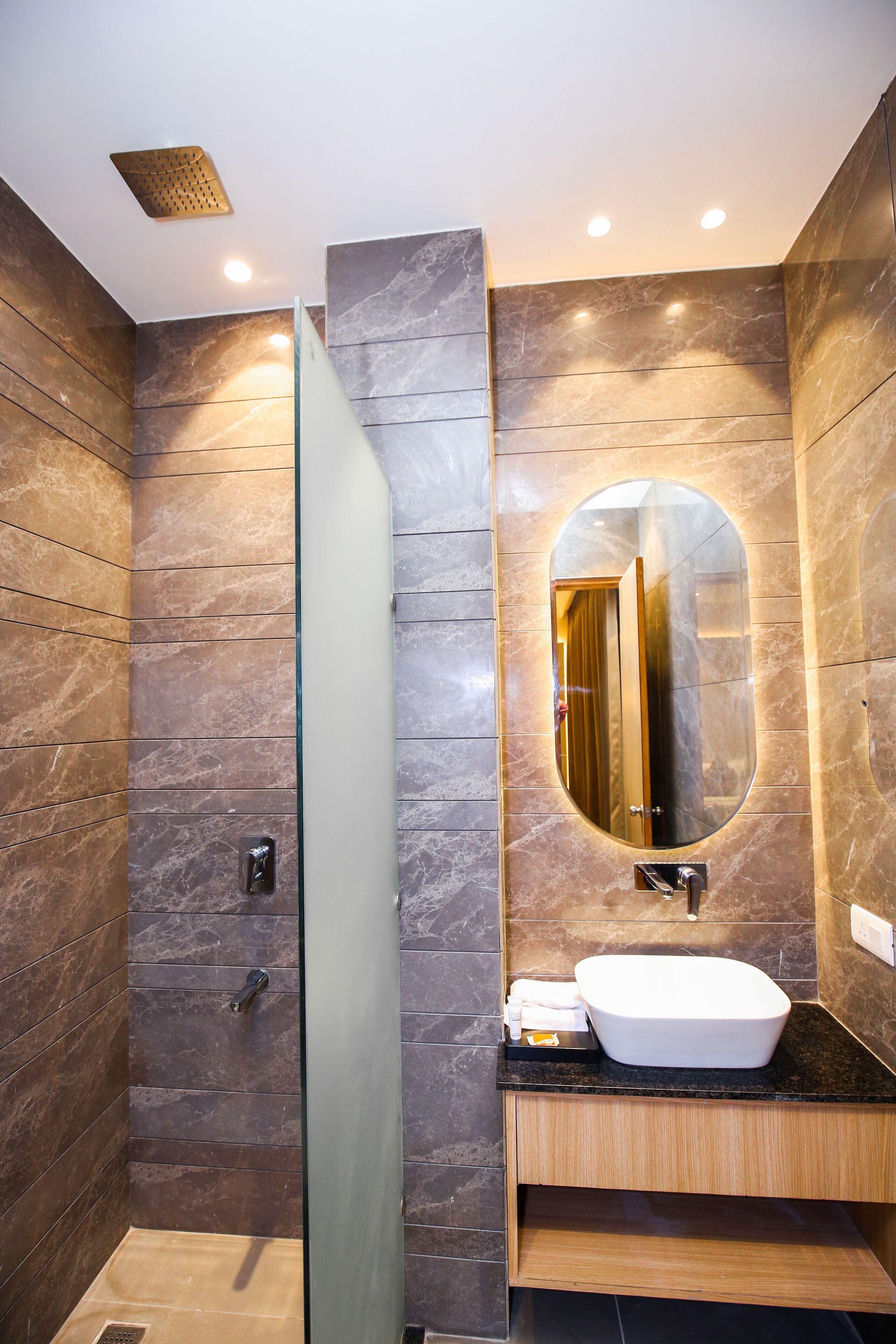 A modern bathroom with a glass shower enclosure, a marble-tiled wall, a wooden vanity, and a large oval mirror.