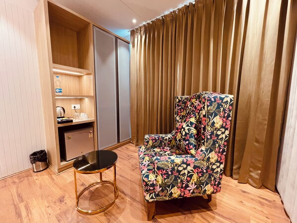 A room with a floral-patterned armchair, a small round table, a kitchenette with a coffee maker, and a wardrobe.