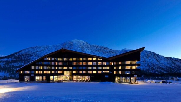 A modern hotel with a symmetrical facade, large windows, and a flat roof, set against a snowy mountain backdrop.