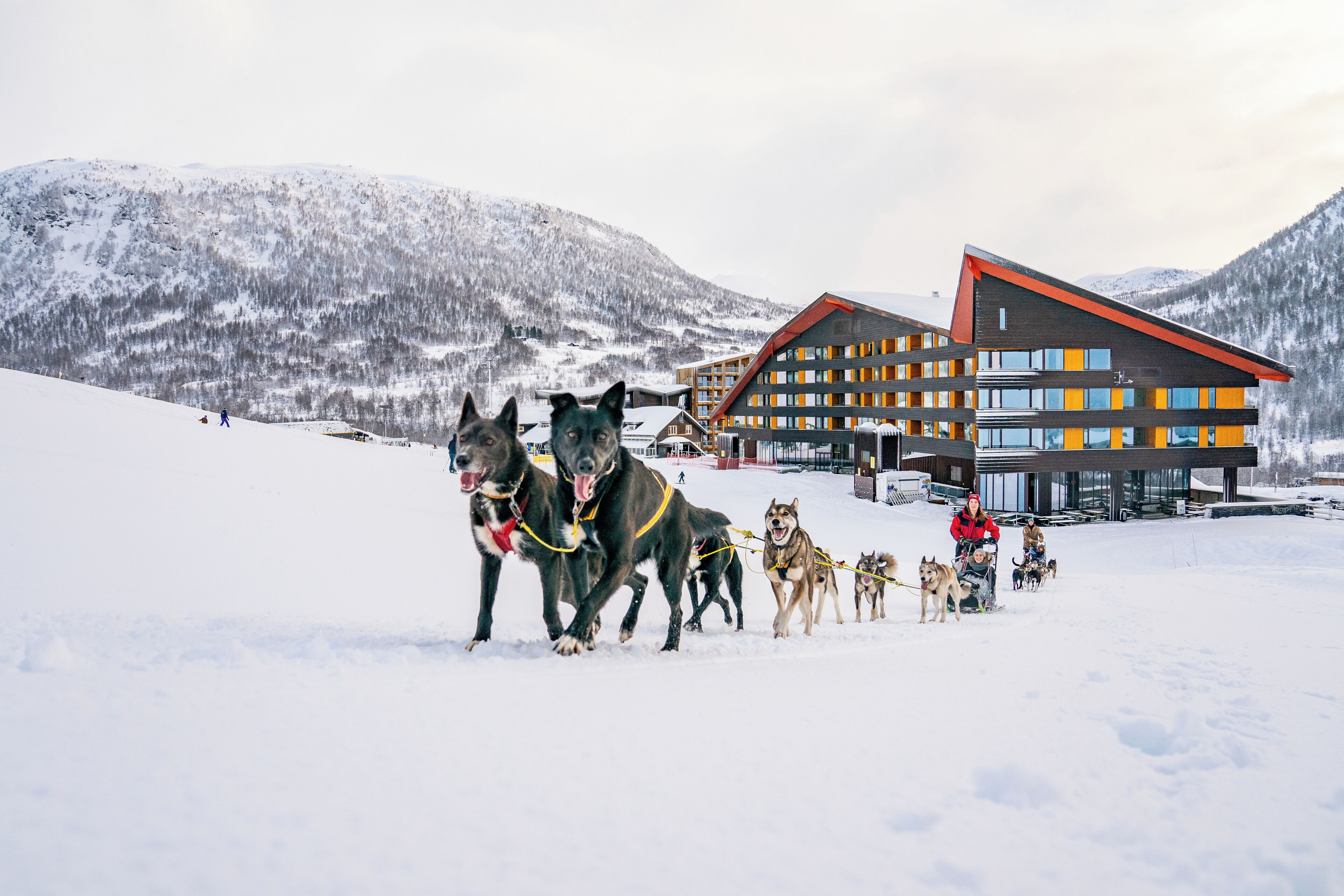 Un edificio moderno con tejado rojo y detalles en amarillo, rodeado de montañas nevadas.