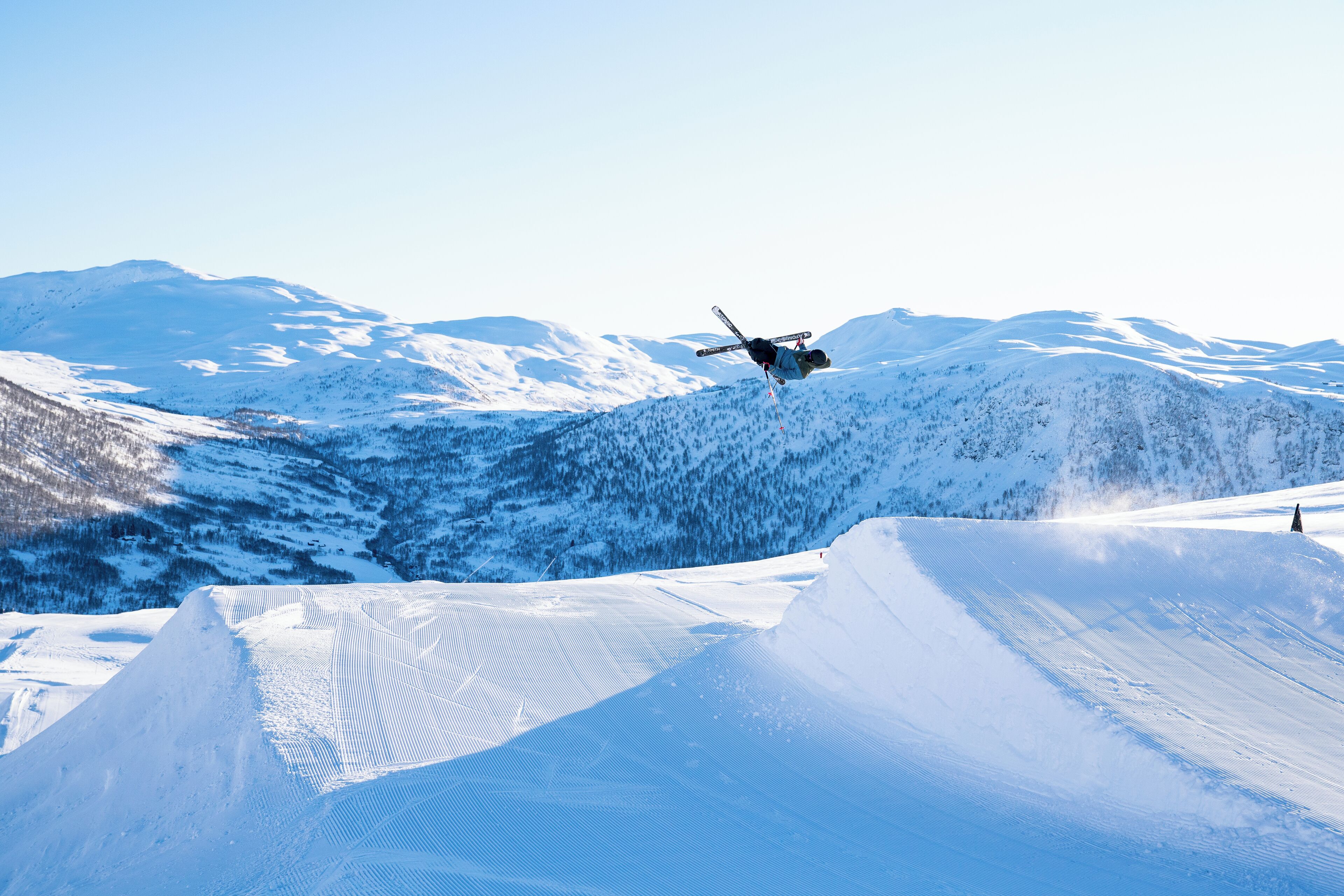 A ski jumper in mid-air over a snow-covered slope with mountains in the background.