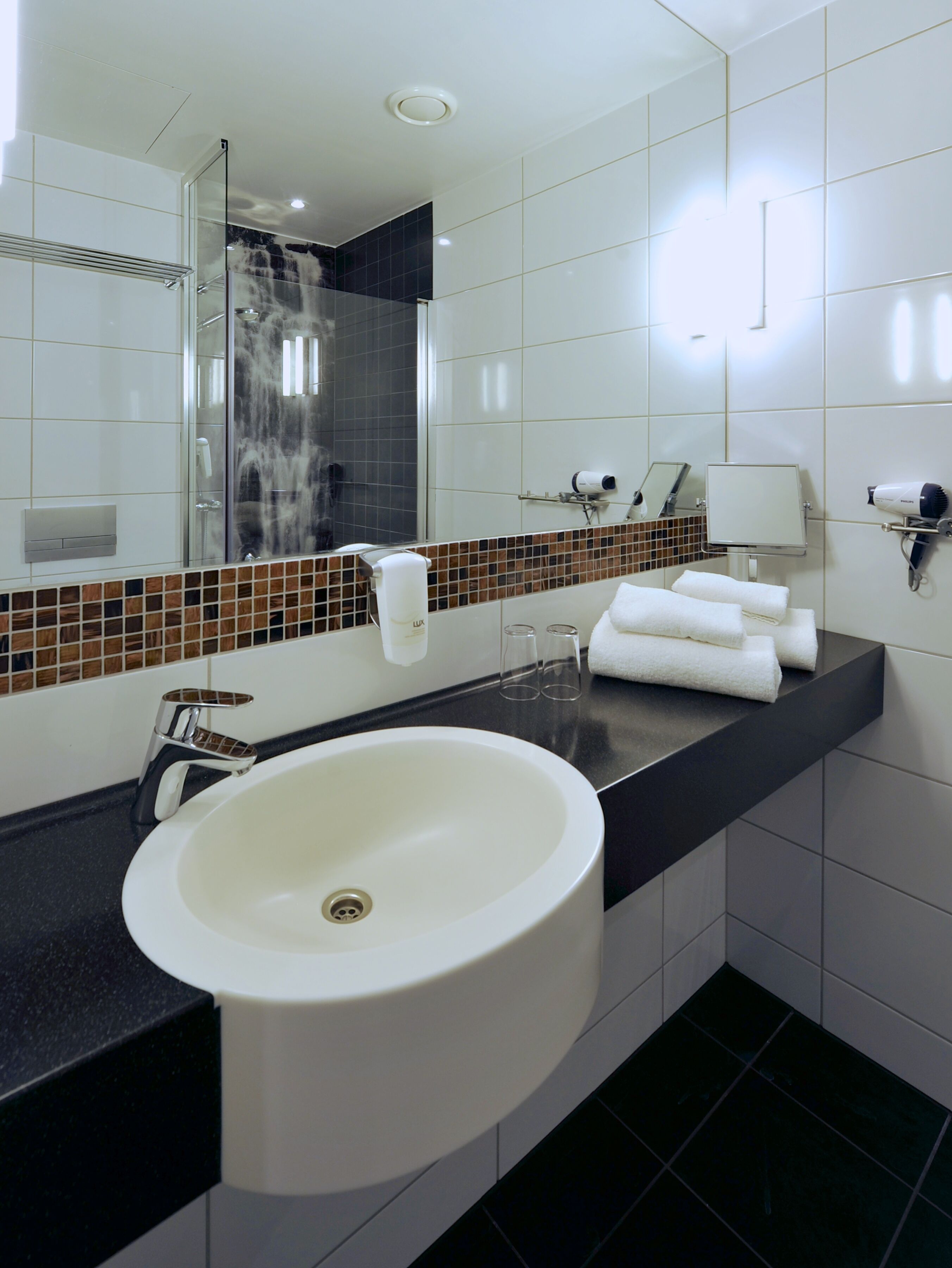 Un baño moderno con un lavabo blanco grande, un espejo, una ducha y un secador de pelo en la encimera.
