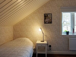 A bedroom with a sloped ceiling, a bed, a nightstand with a lamp, a window with a view, and a floral painting on the wall. - 4 Star Holiday Home in Grebbestad (Grebbestad)