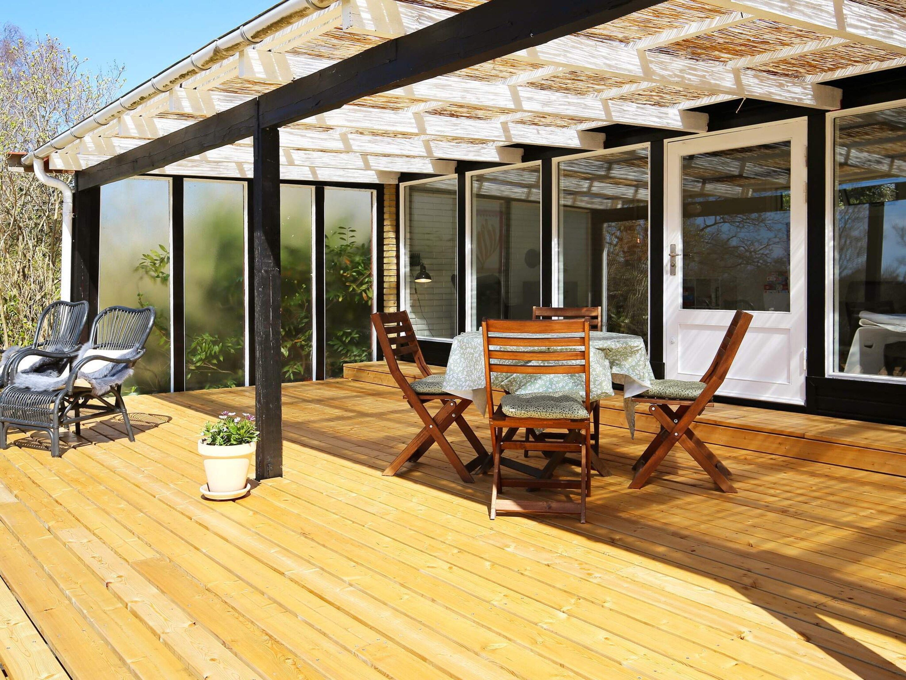 Une terrasse en bois, aménagée avec du mobilier d’extérieur, notamment des chaises et une table, et située sous une zone abritée dotée de grandes fenêtres.