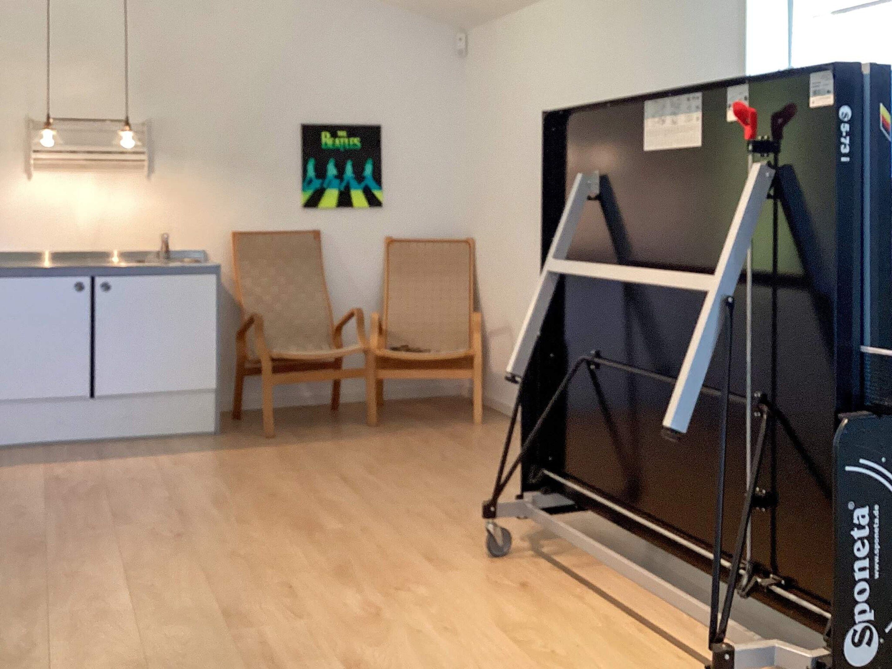 A room with a kitchenette, two wooden chairs, a painting on the wall, and a large black object that appears to be a piece of equipment.