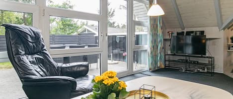 A room with a large window, a black leather chair, a table with a vase of flowers, and a television on a stand.