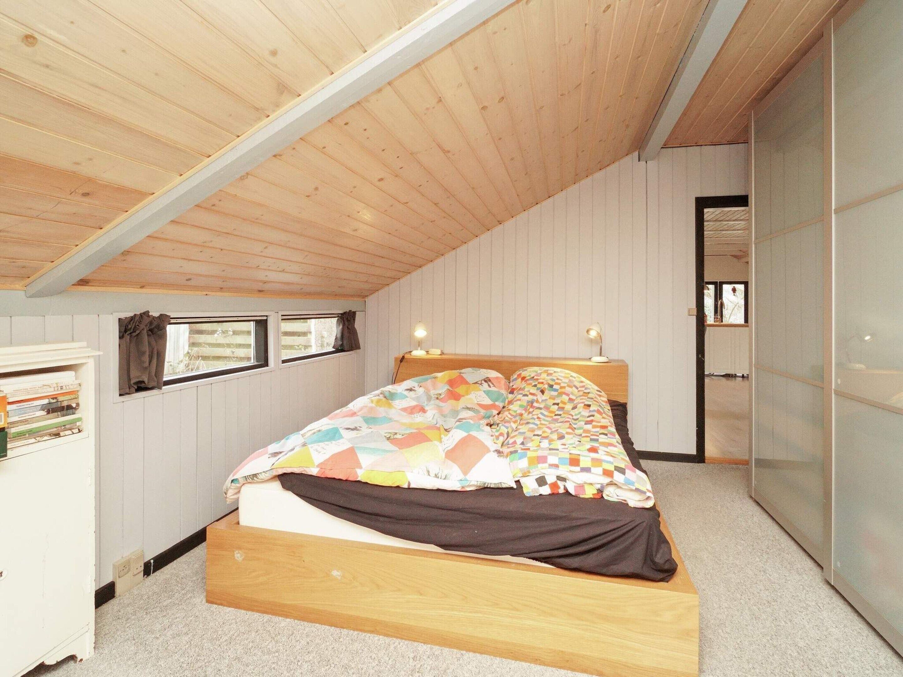 A bedroom with a sloped ceiling, a bed with a colorful quilt, a bedside table with a lamp, and a window with curtains.