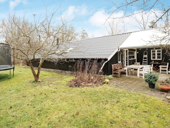 A rustic wooden house with a tiled roof, a small garden with a tree, and outdoor furniture.