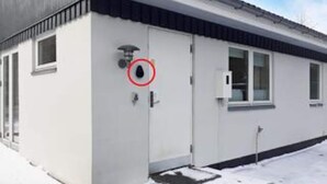 Un edificio con fachada blanca, una puerta con cámara de seguridad y un ventanal.