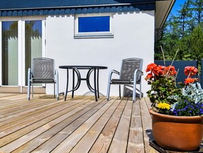 A wooden deck with a table and two chairs, a potted plant with flowers, and a building with a window and a door. - 4 Star Holiday Home in Hojslev (Hojslev)